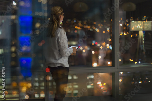 Pensive businesswoman with digital tablet working late, looking out urban office window at night