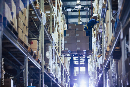 Worker operating forklift stacking cardboard boxes on distribution warehouse shelves