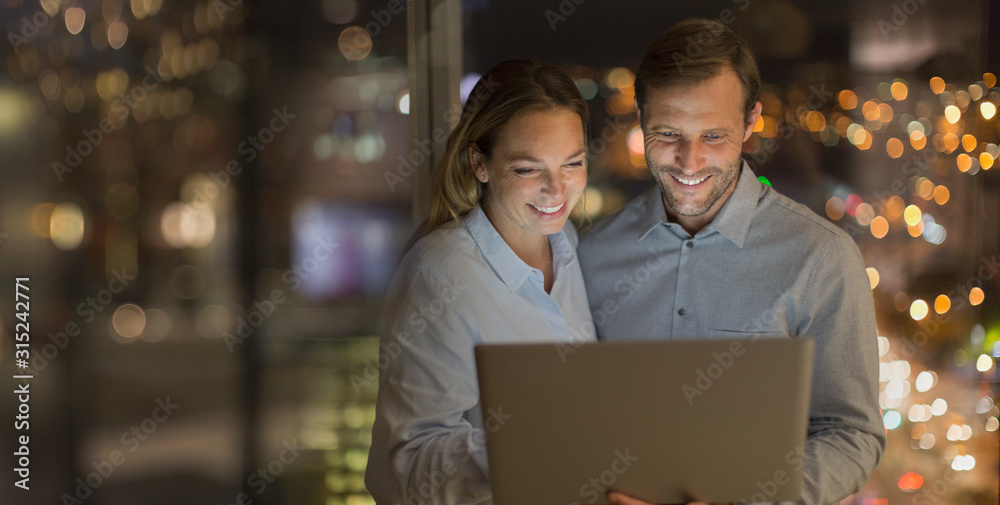 Smiling businessman and businesswoman working late at laptop in office at night Smiling businessman and businesswoman working late at laptop in office at night