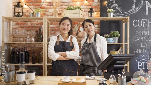 two young confident coffee shop owner women standing in counter and face camera smiling. elegant female barista wear apron joyful in modern cafe bar. Small business entrepreneur together concept.