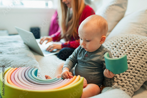 Curious baby girl playing with toy on bed while mother works at laptop photo