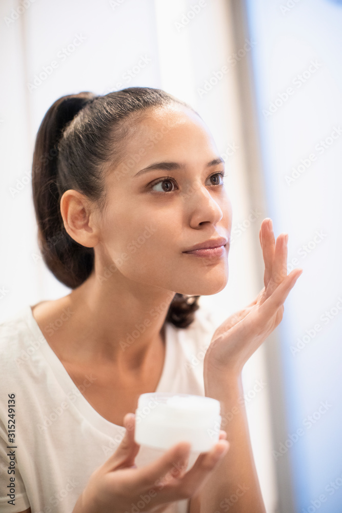 Woman applying moisturizer to face in mirror Woman applying moisturizer to face in mirror