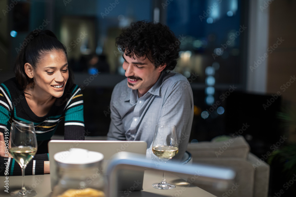 Smiling couple using laptop and drinking white wine at home at night Smiling couple using laptop and drinking white wine at home at night