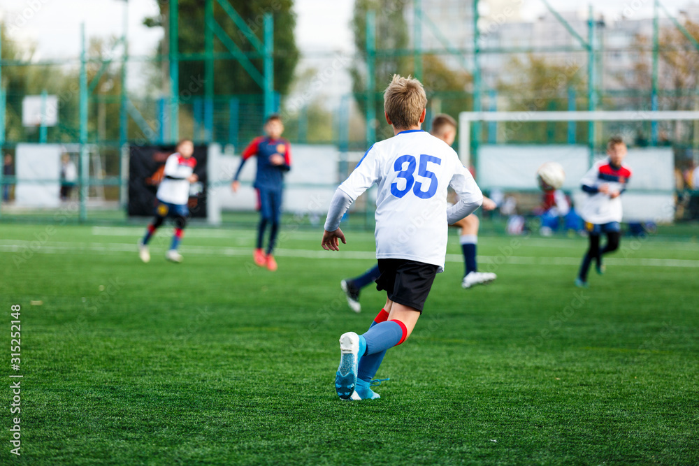 Boys in white and blue sportswear plays football on field, dribbles ball. Young soccer players with ball on green grass. Training, football, active lifestyle for kids concept