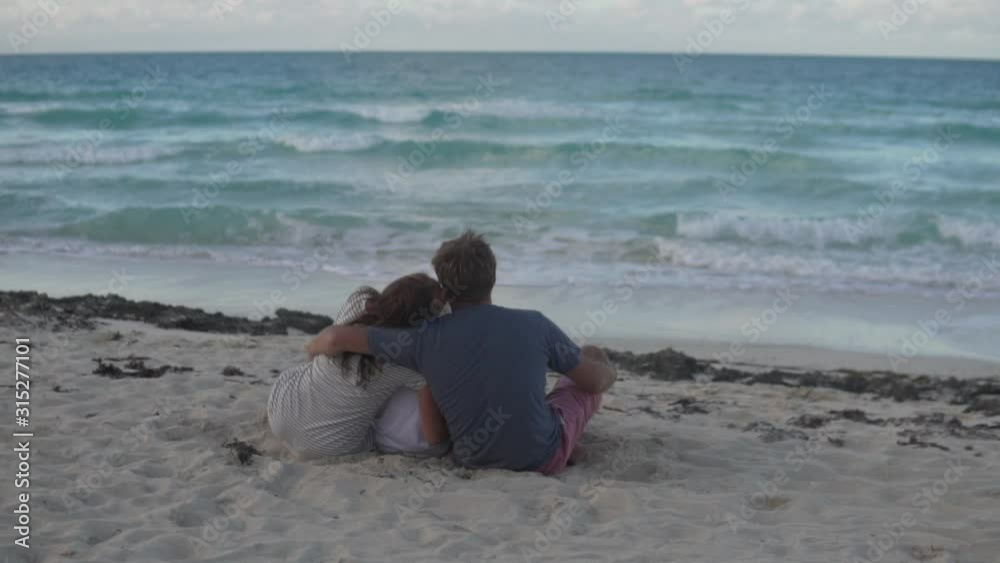 The family sits with their backs to the camera on the beach by the ocean. Parents hug each other and the blonde's son The family sits with their backs to the camera on the beach by the ocean. Parents hug each other and the blonde's son