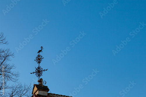 Background image - a metal weather vane in the shape of a stork sitting on a base supported by a stone lion against a clear blue sky