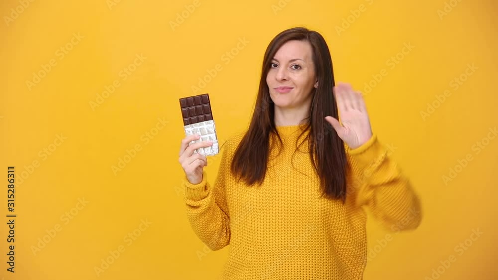 Young woman in bright yellow sweater shirt posing hold in hand chocolate bar say no showing stop gesture isolated over yellow orange background in studio. People sincere emotions lifestyle concept. Young woman in bright yellow sweater shirt posing hold in hand chocolate bar say no showing stop gesture isolated over yellow orange background in studio. People sincere emotions lifestyle concept.