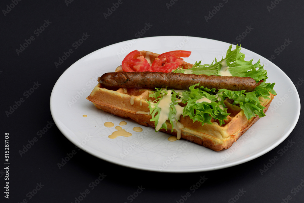 Classic sausage on waffles breakfast with tomatoes, salad and sauce served on a white plate on black background. Classic sausage on waffles breakfast with tomatoes, salad and sauce served on a white plate on black background.
