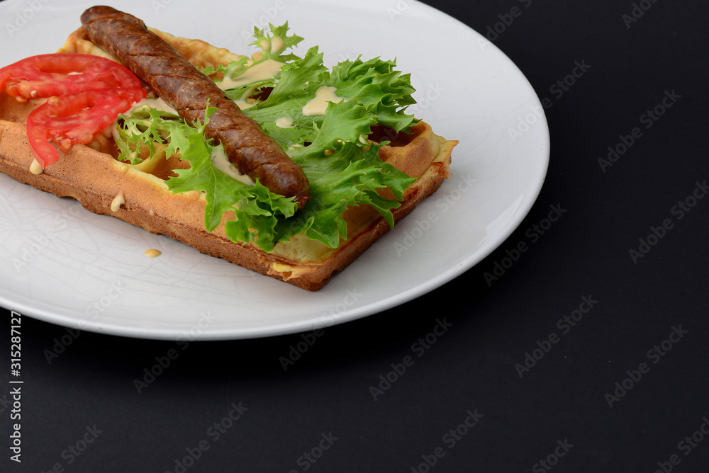 Classic sausage on waffles breakfast with tomatoes, salad and sauce served on a white plate on black background.