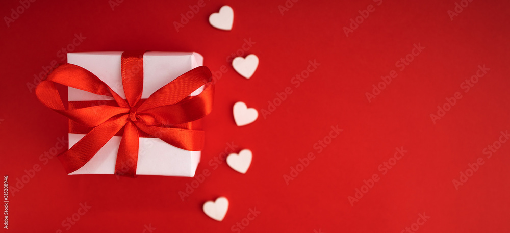 Top view of present box package decorated into the white paper and red bow-knot with signs of white wooden hearts besides. Flat lay on red background. Concept of celebrate. Top view of present box package decorated into the white paper and red bow-knot with signs of white wooden hearts besides. Flat lay on red background. Concept of celebrate.