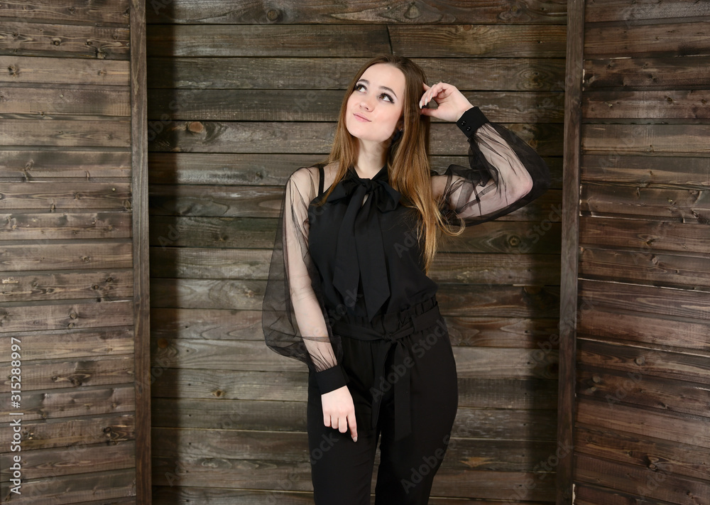 The concept of home comfort. Photo of a pretty brunette girl with excellent make-up in dark clothes standing on a wooden background in the home interior, smiling. The concept of home comfort. Photo of a pretty brunette girl with excellent make-up in dark clothes standing on a wooden background in the home interior, smiling.