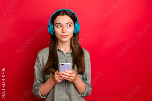 Photo of youngster lady using modern technology headphones listen song holding telephone dreamy look empty space wear casual grey green shirt isolated red color background