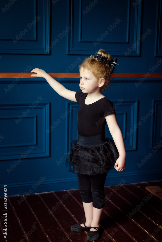 View of little blonde curly ballerina in black leotard practicing at ballet barre stand one at dance studio. Kids hobby dancing. Ballet is hard work for children. Active lifestyle in childhood View of little blonde curly ballerina in black leotard practicing at ballet barre stand one at dance studio. Kids hobby dancing. Ballet is hard work for children. Active lifestyle in childhood