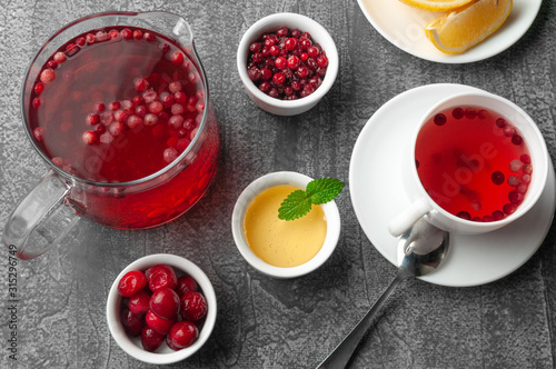 Glass jug with a drink of cranberries and cherries. Next to the jug are plates with berries, honey and a white porcelain cup. View from above. On a gray concrete background.