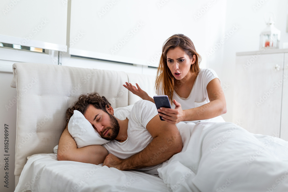 Jealous wife spying the phone of her partner while he is sleeping in a bed at home. Shocked