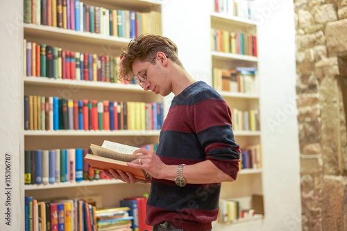 Bild auf Leinwand Young student with a watch and glasses, reading a book in library next to a bookshelf