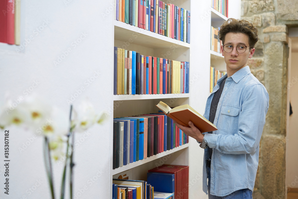 Student with glasses and book in his hand looking to the side. Stone wall in the background. Flower in the foreground. Student with glasses and book in his hand looking to the side. Stone wall in the background. Flower in the foreground.