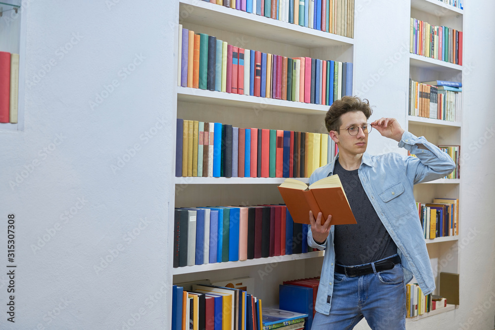 Young student next to bookshelf with book in hand checking his glasses