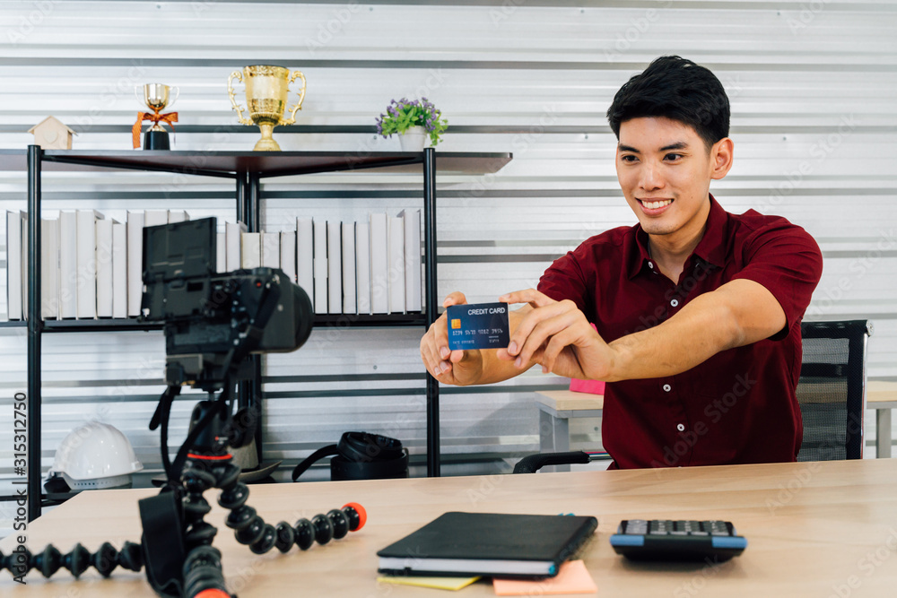Smiling Asian male recording video selling stuff through social media and video platform showing credit card using camera sitting at table in house