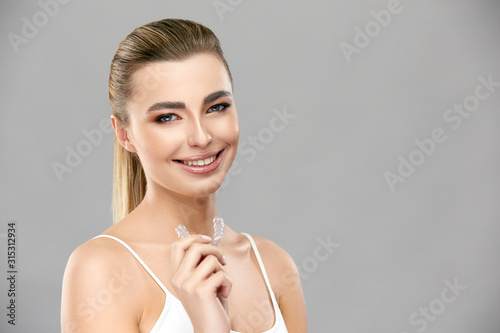 pretty girl with perfect smile holding invisible braces and smiling to camera on grey background