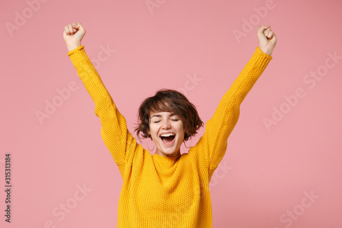 Happy young brunette woman girl in yellow sweater posing isolated on pastel pink background studio portrait. People lifestyle concept. Mock up copy space. Doing winner gesture, keeping eyes closed. photo