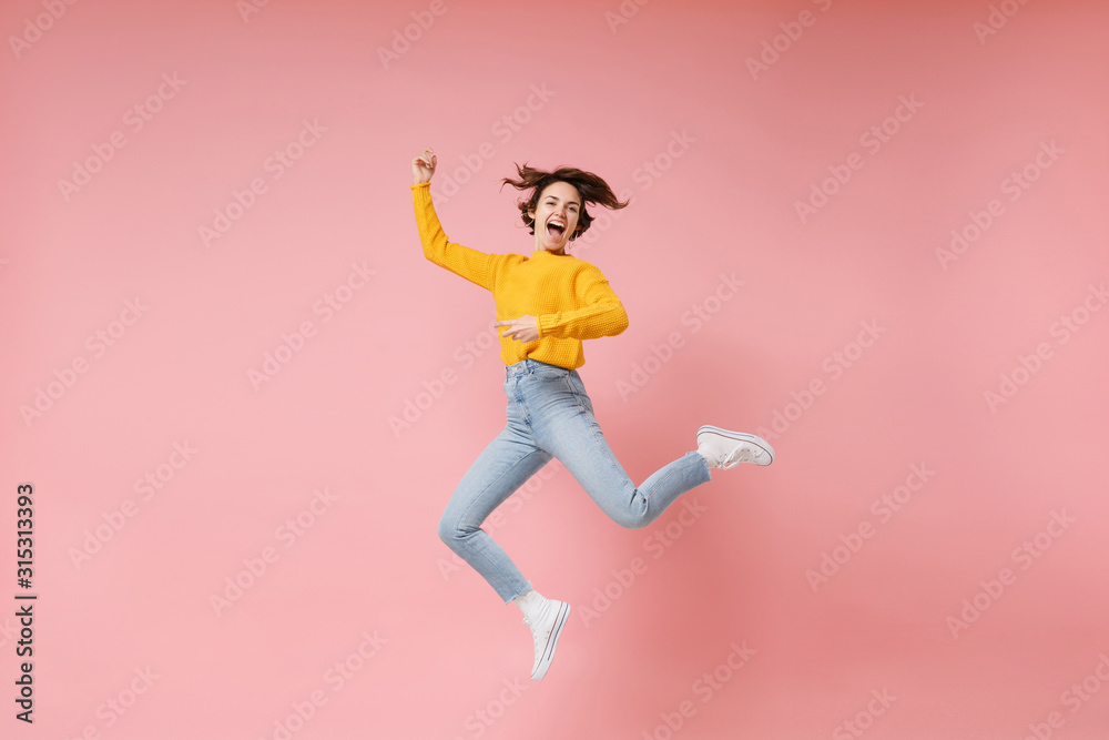 Excited young brunette woman girl in yellow sweater posing isolated on pastel pink background in studio. People lifestyle concept. Mock up copy space. Having fun fooling around, rising hands, jumping.