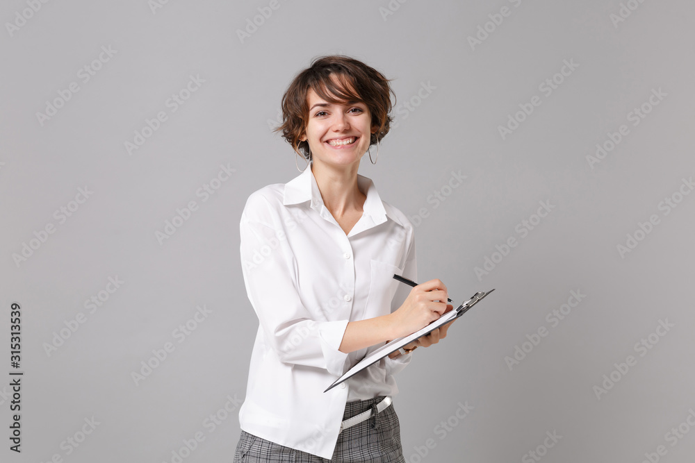 Fototapeta premium Smiling young business woman in white shirt posing isolated on grey wall background. Achievement career wealth business concept. Mock up copy space. Hold clipboard with papers document, writing notes.