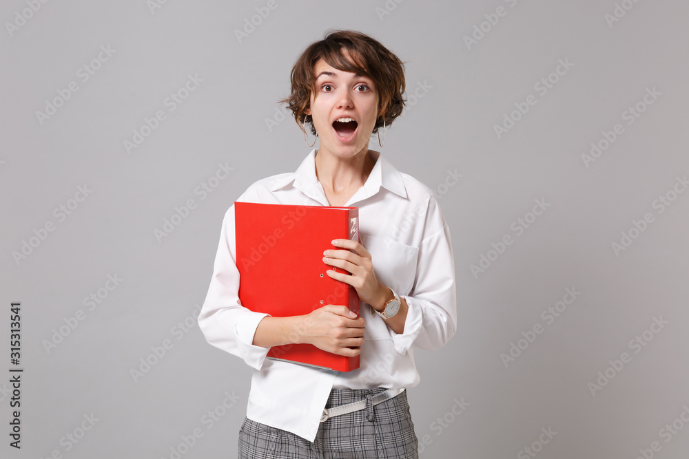 Shocked young business woman in white shirt posing isolated on grey background studio portrait. Achievement career wealth business concept. Mock up copy space. Holding red folder for papers document. Shocked young business woman in white shirt posing isolated on grey background studio portrait. Achievement career wealth business concept. Mock up copy space. Holding red folder for papers document.
