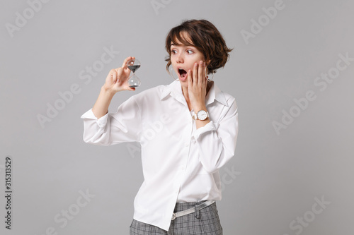 Shocked young business woman in white shirt posing isolated on grey wall background studio portrait. Achievement career wealth business concept. Mock up copy space. Hold hourglass, put hand on cheek.