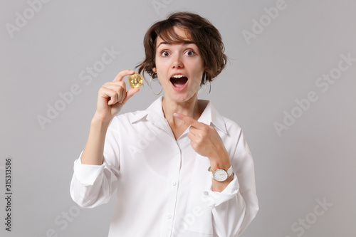 Excited young business woman in white shirt posing isolated on grey wall background. Achievement career wealth business concept. Mock up copy space. Poiting index finger on bitcoin, future currency.
