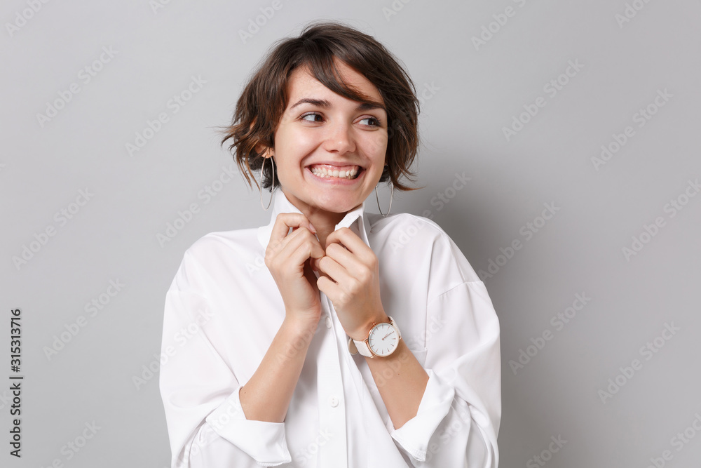 Smiling young business woman in white shirt posing isolated on grey wall background studio portrait. Achievement career wealth business concept. Mock up copy space. Straightening collar looking aside. Smiling young business woman in white shirt posing isolated on grey wall background studio portrait. Achievement career wealth business concept. Mock up copy space. Straightening collar looking aside.
