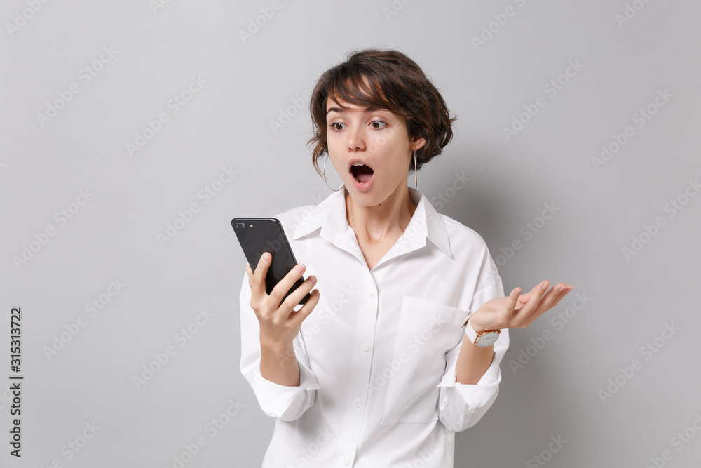 Amazed young business woman in white shirt posing isolated on grey background studio portrait. Achievement career wealth business concept. Mock up copy space. Holding mobile phone, spreading hands. Amazed young business woman in white shirt posing isolated on grey background studio portrait. Achievement career wealth business concept. Mock up copy space. Holding mobile phone, spreading hands.