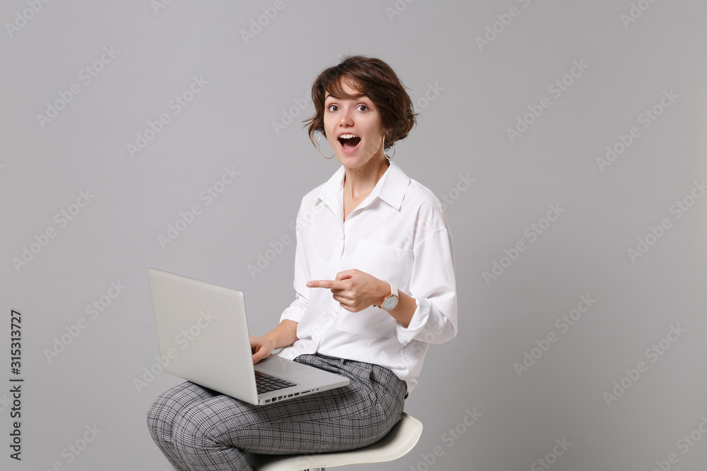 Excited young business woman in white shirt isolated on grey background in studio. Achievement career wealth business concept. Mock up copy space. Pointing index finger on laptop pc computer, sitting. Excited young business woman in white shirt isolated on grey background in studio. Achievement career wealth business concept. Mock up copy space. Pointing index finger on laptop pc computer, sitting.