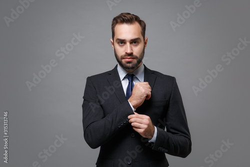Successful young business man in classic black suit shirt tie posing isolated on grey background studio portrait. Achievement career wealth business concept. Mock up copy space. Straightening sleeves.