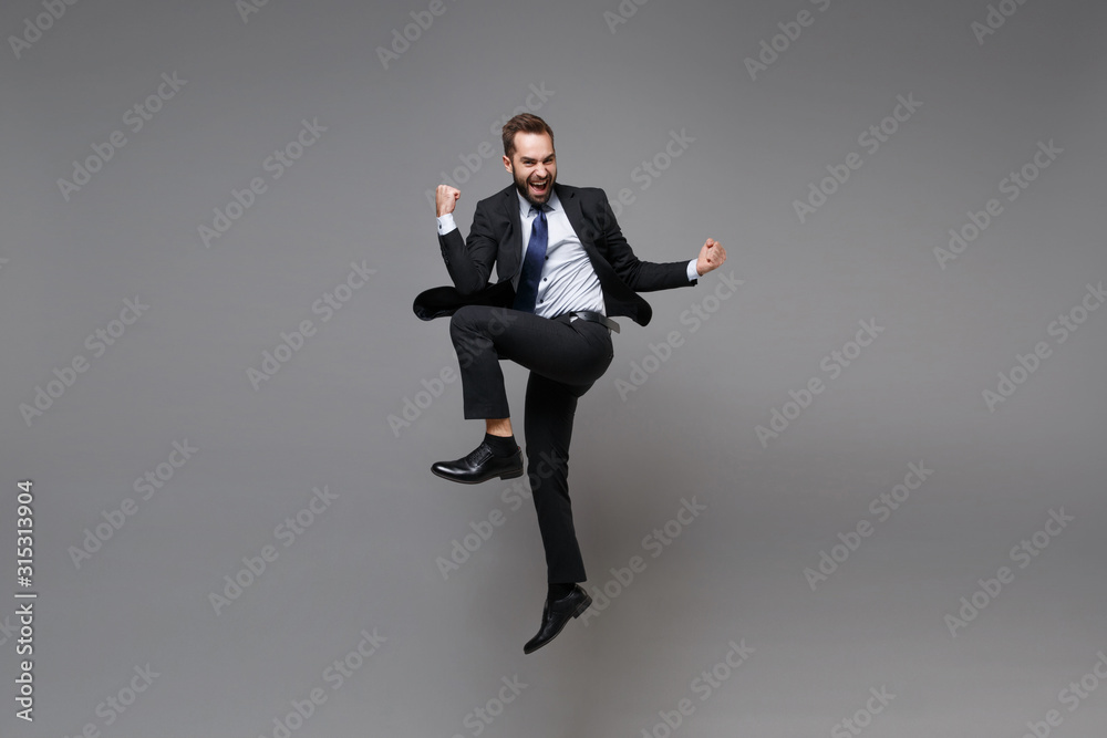 Overjoyed young business man in classic black suit shirt tie posing isolated on grey background. Achievement career wealth business concept. Mock up copy space. Jumping doing winner gesture screaming. Overjoyed young business man in classic black suit shirt tie posing isolated on grey background. Achievement career wealth business concept. Mock up copy space. Jumping doing winner gesture screaming.