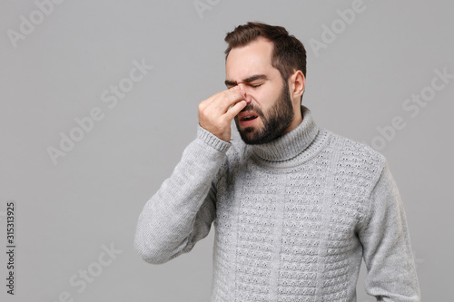 Young man in gray sweater posing isolated on grey background in studio. Healthy lifestyle, ill sick disease treatment, cold season concept. Mock up copy space. Put hand on nose keeping eyes closed.