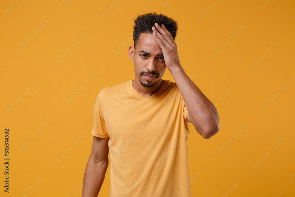 Exhausted young african american guy in casual t-shirt posing isolated on yellow orange wall background studio portrait. People emotions lifestyle concept. Mock up copy space. Putting hand on head. Exhausted young african american guy in casual t-shirt posing isolated on yellow orange wall background studio portrait. People emotions lifestyle concept. Mock up copy space. Putting hand on head.