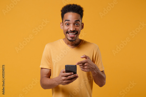 Funny young african american guy in casual t-shirt posing isolated on yellow orange background, studio portrait. People lifestyle concept. Mock up copy space. Pointing index finger on mobile phone.