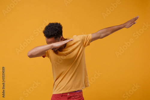 Young african american guy in casual t-shirt posing isolated on yellow orange wall background studio portrait. People sincere emotions lifestyle concept. Mock up copy space. Showing DAB dance gesture. photo