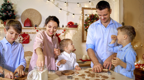 Whole family is making gingerbread cookies together for Christmas at xmas eve. Family of five people is preparing a dough for cookie at kitchen with xmas decoration.