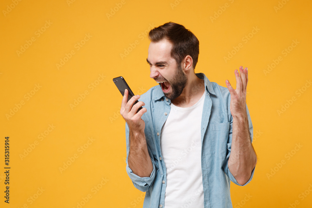 Angry young man in casual blue shirt posing isolated on yellow orange wall background studio portrait. People sincere emotions lifestyle concept. Mock up copy space. Screaming talking on mobile phone. Angry young man in casual blue shirt posing isolated on yellow orange wall background studio portrait. People sincere emotions lifestyle concept. Mock up copy space. Screaming talking on mobile phone.