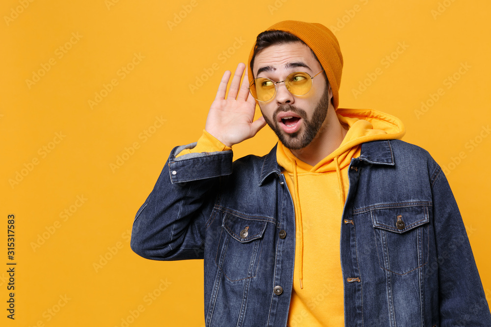 Curious young hipster guy in fashion jeans denim clothes posing isolated on yellow orange background studio portrait. People lifestyle concept. Mock up copy space. Try to hear you with hand near ear. Curious young hipster guy in fashion jeans denim clothes posing isolated on yellow orange background studio portrait. People lifestyle concept. Mock up copy space. Try to hear you with hand near ear.