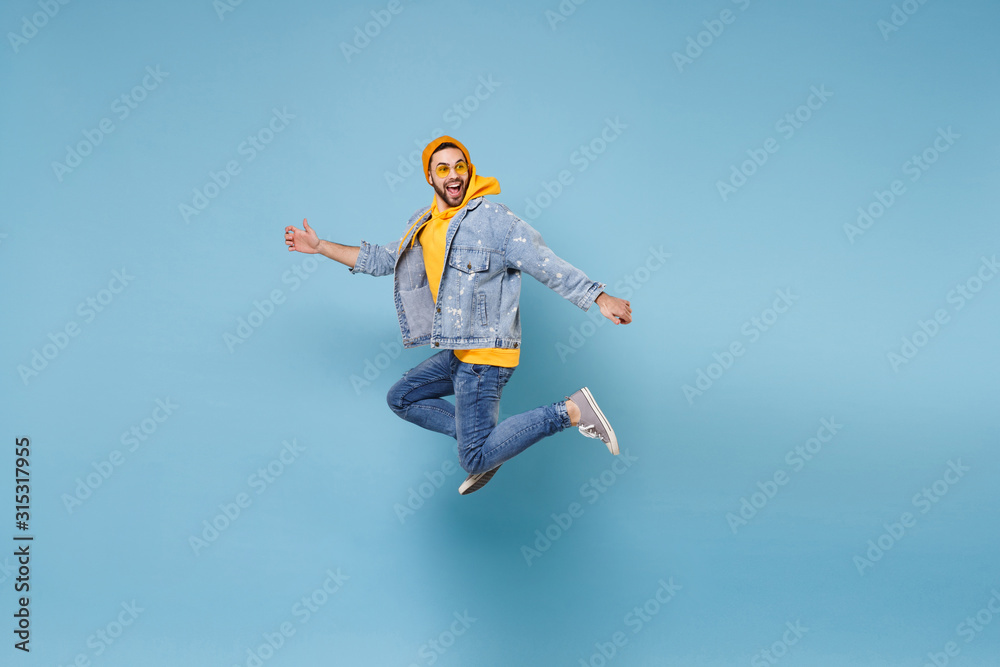 Obraz premium Side view of funny young hipster guy in fashion jeans denim clothes posing isolated on pastel blue background. People lifestyle concept. Mock up copy space. Jumping, spreading hands, looking aside.