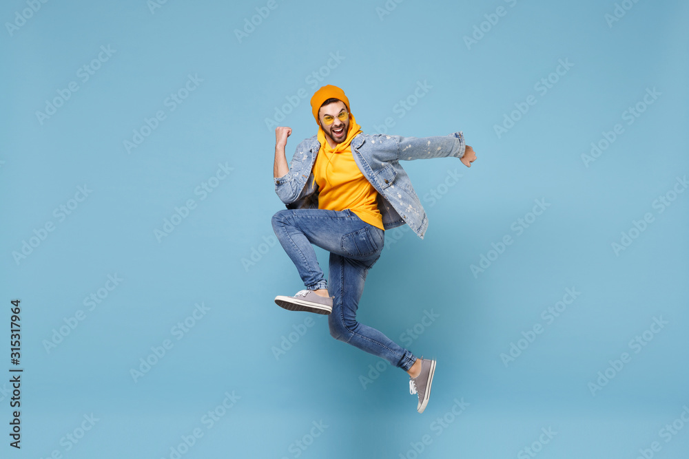 Side view of happy young hipster guy in fashion jeans denim clothes posing isolated on pastel blue background studio portrait. People lifestyle concept. Mock up copy space. Jump doing winner gesture. - obrazy, fototapety, plakaty 