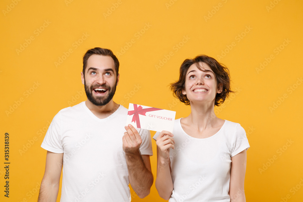 Smiling young couple friends bearded guy girl in white blank empty t-shirts isolated on yellow orange wall background. People lifestyle concept. Mock up copy space. Hold gift certificate, looking up. Smiling young couple friends bearded guy girl in white blank empty t-shirts isolated on yellow orange wall background. People lifestyle concept. Mock up copy space. Hold gift certificate, looking up.