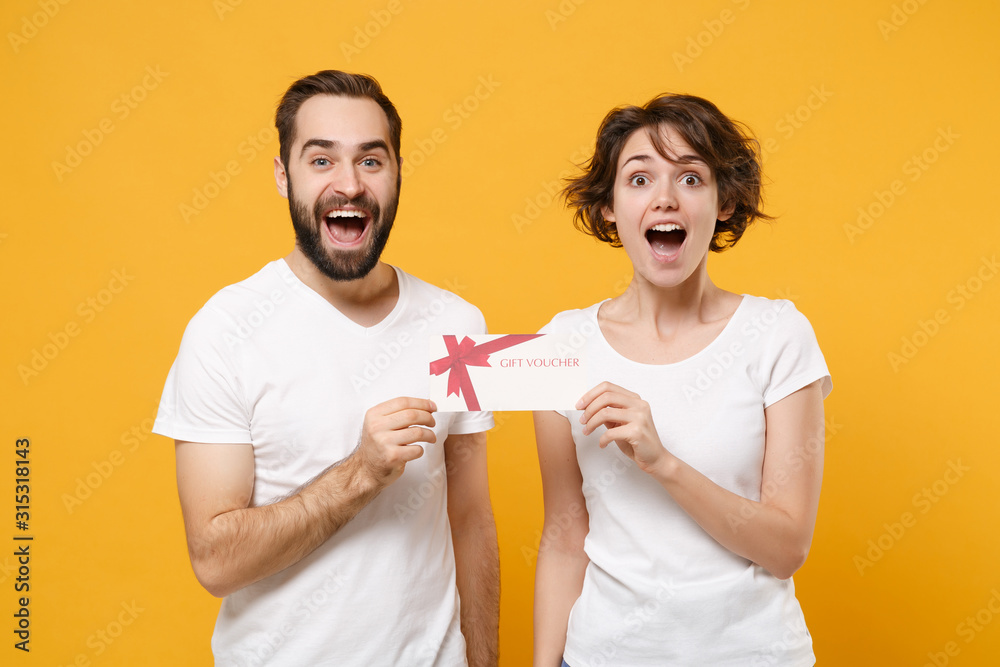 Excited young couple friends bearded guy girl in white blank empty t-shirts isolated on yellow orange wall background in studio. People lifestyle concept. Mock up copy space. Holding gift certificate. Excited young couple friends bearded guy girl in white blank empty t-shirts isolated on yellow orange wall background in studio. People lifestyle concept. Mock up copy space. Holding gift certificate.