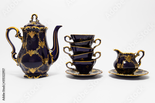 Closeup of a beautiful cobalt blue colored vintage porcelain tea set with golden floral pattern on white background. The set includes a tea pot, a milk jug and a stack of tea cups.