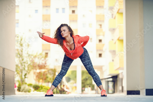 Fototapeta Naklejka Na Ścianę i Meble -  Focused attractive caucasian woman in sportswear and with curly hair bending with arm stretched and doing warm up exercises while standing wide-legged in passage on sunny day.