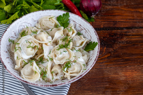 Boiled dumplings with feathers of green onions. In the background are greens, red peppers and bay leaves. In a transparent bowl sour cream.