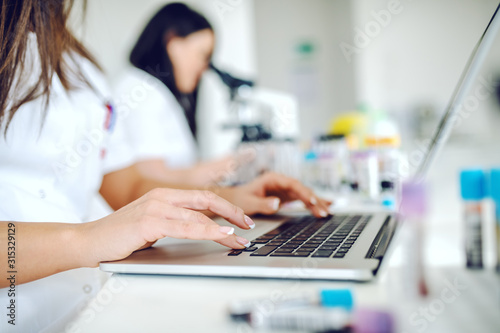Close up of female lab assistant in white uniform sitting in lab and using laptop for data entry. In background is her colleague working.